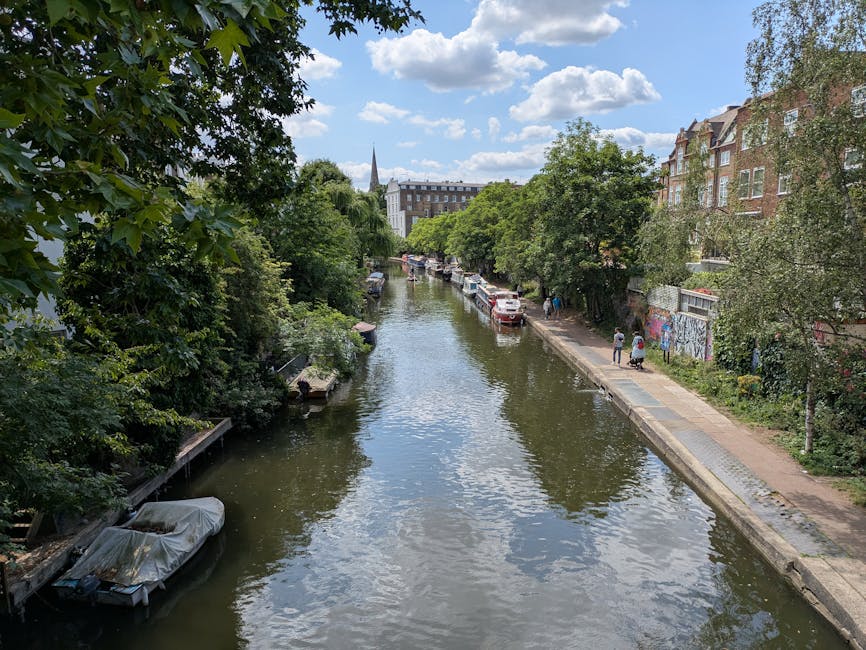 A canal scene during daytime in Islington showing a narrow waterway lined with green trees on both sides, with several boats moored along the edges, some covered with protective tarps. On the right side of the canal, there is a pedestrian pathway with a few people walking, and cars parked along the street. On the left side, lush foliage extends over the water, partially obscuring the boats. In the background, residential buildings and a church spire are visible under a partly cloudy sky. The overall scene reflects an urban environment suitable for house removals and furniture transport, with visible elements for loading and moving logistics, supported by companies like Man with Van Canonbury.