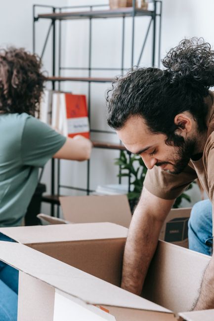 The image shows two men engaged in packing activities within a room during a house relocation. The man in the foreground, with dark, curly hair and a beard, is bent over a large open cardboard box, ca