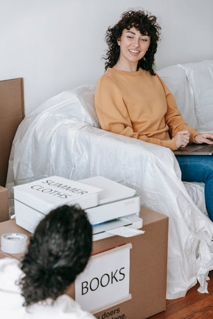 The image shows two men engaged in packing activities within a room during a house relocation. The man in the foreground, with dark, curly hair and a beard, is bent over a large open cardboard box, ca