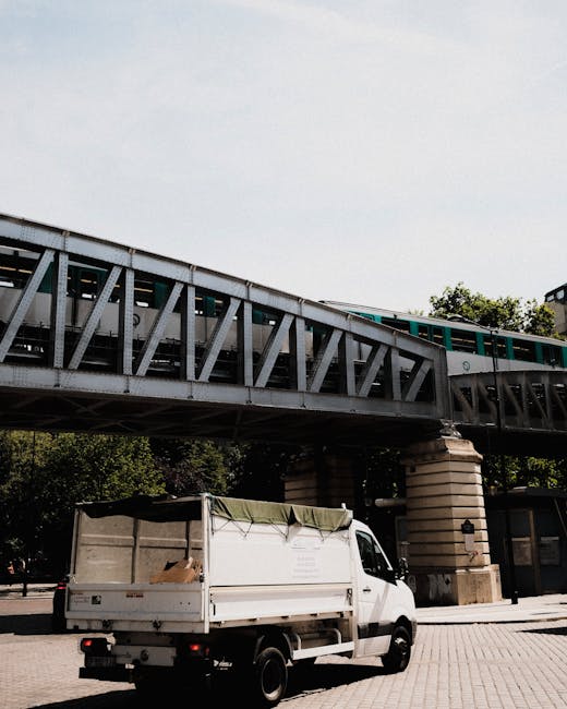 A white commercial van with an open cargo area is parked on a paved area near a low wall, beneath a railway bridge with metal framework and concrete supports. The van's cargo area has a green cover se