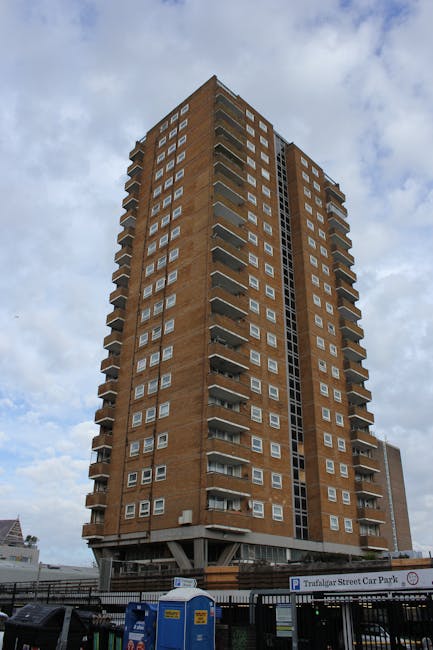 An upward view of a tall, multi-storey residential building constructed with brown brickwork, featuring numerous small rectangular windows aligned in a grid pattern across its façade. The building has several protruding balconies with metal railings on each level, and visible fire escape stairs run vertically along one side. The sky above is partly cloudy with patches of blue. At the base of the building, there is a parking area with a sign indicating Trafalgar Street Car Park, and a black metal fence surrounds the site. In the foreground, there are portable toilets and a white van, suggesting recent moving or building maintenance activity, aligning with professional house removals and furniture transport services provided by Man with Van Canonbury.