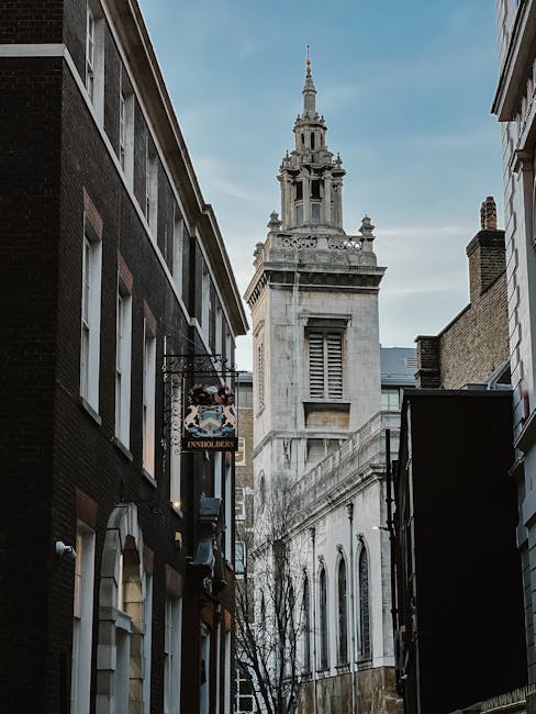 A narrow urban alleyway between two rows of tall buildings leads towards a historic church tower with a spire and ornate architectural details, visible in the background beneath a cloudy sky. On the left side of the alley, there are commercial storefronts with signage, including one for 'Insholders', and street-level windows. The area appears to be part of a city centre with closely spaced structures, possibly during a home relocation or moving process. Although no furniture or packing materials are visible in this image, the scene sets an environment where house removals and furniture transport services like those offered by Man with Van Canonbury could be involved. The lighting is natural, with daylight illuminating the scene, emphasizing the historic and urban context of the property and the moving logistics associated with the area.
