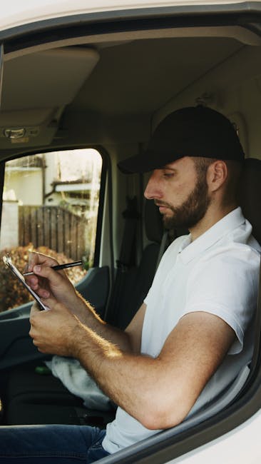 A man wearing a white shirt, blue jeans, and a black cap is seen loading three cardboard boxes into the front of a white delivery van parked on a residential street. The van, which belongs to Man with