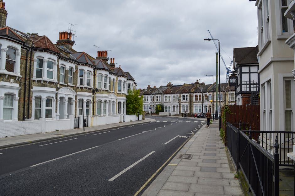 A residential street scene showing a row of Victorian-style terraced houses with bay windows and brick facades, some painted white, on the left side of the image. The sidewalk on the right features a black metal railing, with a wooden fence supporting climbing plants. A man wearing dark clothing is walking along the sidewalk, while a few parked cars are visible further down the street. The road is asphalt with clear white lane markings, slightly curved, and lined with street lamps and trees. The sky is overcast with gray clouds, indicating typical weather during a home relocation or furniture transport process. This scene depicts an urban area suitable for house removals and moving services such as those provided by Man with Van Canonbury, with the environment prepared for moving logistics and transportation.