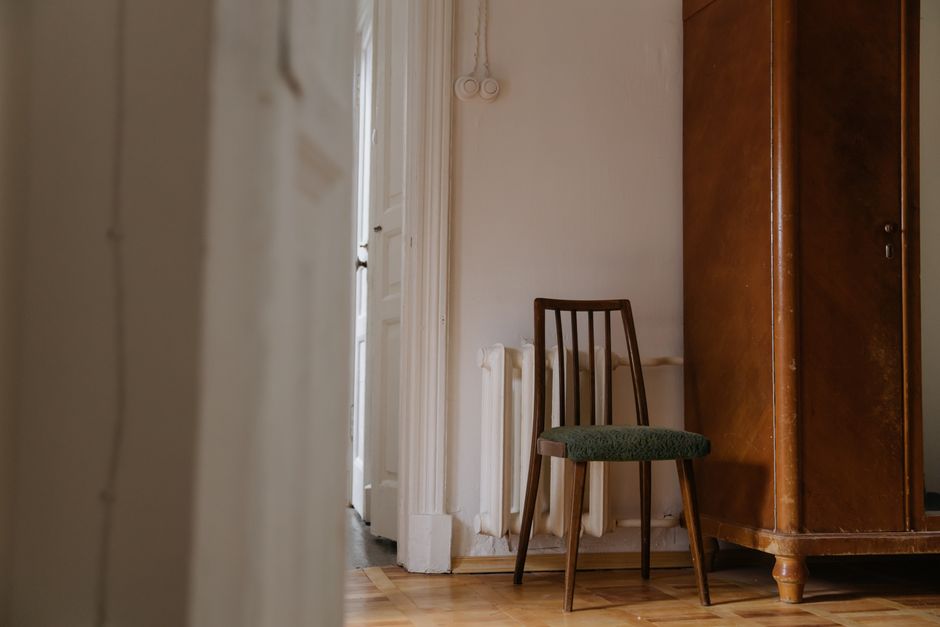 A small wooden chair with a green upholstered seat positioned inside a room near a white internal door with glass panels, revealing a narrow passageway or doorway outside. To the right of the chair is a tall wooden wardrobe with a reddish-brown finish, partially blocking the view. The room has light-colored walls and a wooden parquet floor. The scene depicts an indoor environment during a home relocation or packing process, with natural lighting illuminating the space. The arrangement suggests the furniture is in the process of being moved, possibly by a professional removals service such as Man with Van Canonbury, who assist with furniture transport and fitting difficult items through doorways during house removals.