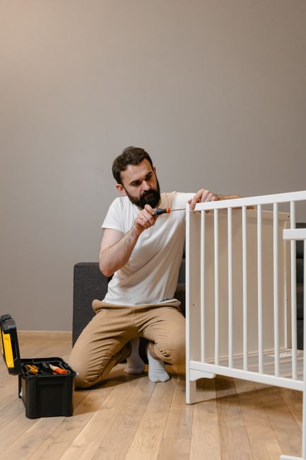 A man with a beard, wearing a white t-shirt and beige trousers, is kneeling on a wooden floor next to a white baby cot, using a screwdriver to assemble or adjust the cot's frame. There is a black toolbox with a yellow handle open on the floor nearby, containing various tools. The background features a plain, neutral-colored wall and a gray sofa against the wall. This scene appears to be set inside a home during a furniture assembly or home relocation process, with natural indoor lighting illuminating the workspace. The man is focused on the task, supporting the furniture's stability, which is part of the house move or furniture fitting service offered by Man with Van Canonbury, involved in furniture transport and packing for home relocations.