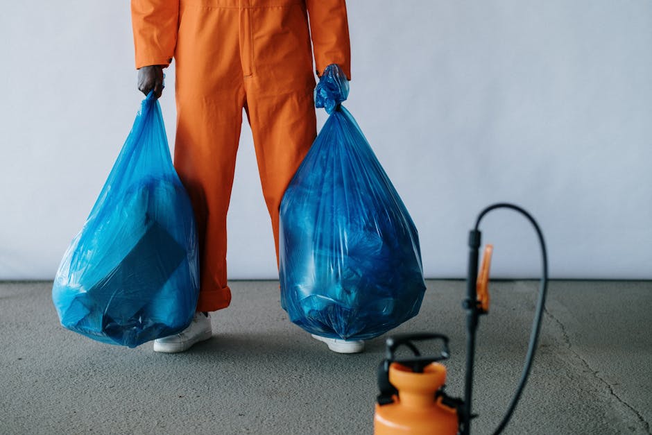 A person dressed in orange work overalls is standing indoors on a concrete floor, holding two large blue plastic trash bags filled with waste or recyclable materials, with one bag in each hand. The individual's face is not visible, only their legs and hands. Nearby, an orange vacuum-type machine with a black cord and a spray nozzle attachment is placed on the floor. The background features a plain, light-colored wall, suggesting an indoor setting related to home responsibilities such as packing or disposal. This scene depicts a typical process involved in home relocation or removal services, with an emphasis on waste collection and packing for moving, which aligns with house removal activities and the logistical aspects of furniture transport and packing.