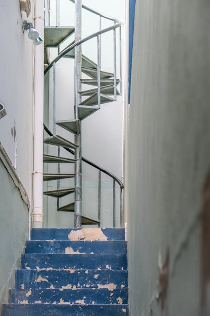 Inside a narrow, old Victorian staircase featuring worn blue-painted wooden steps with chipped paint revealing the bare wood beneath. The staircase is enclosed by plain white walls and, at the top, a metallic spiral staircase with a silver handrail, likely made of metal or steel, is visible. To the left, a white pipe runs down the wall, alongside a small window that provides natural light into the space. The environment appears to be part of an older residential property, possibly during a house removal process. The photograph was taken from the bottom of the stairs, highlighting the confined space and the transition between the lower and upper levels. This setting illustrates typical challenges of navigating narrow staircases during home relocations, where careful handling of furniture and boxes is essential. Man with Van Canonbury may be involved in such services, focusing on house removals and furniture transport in similar environments.