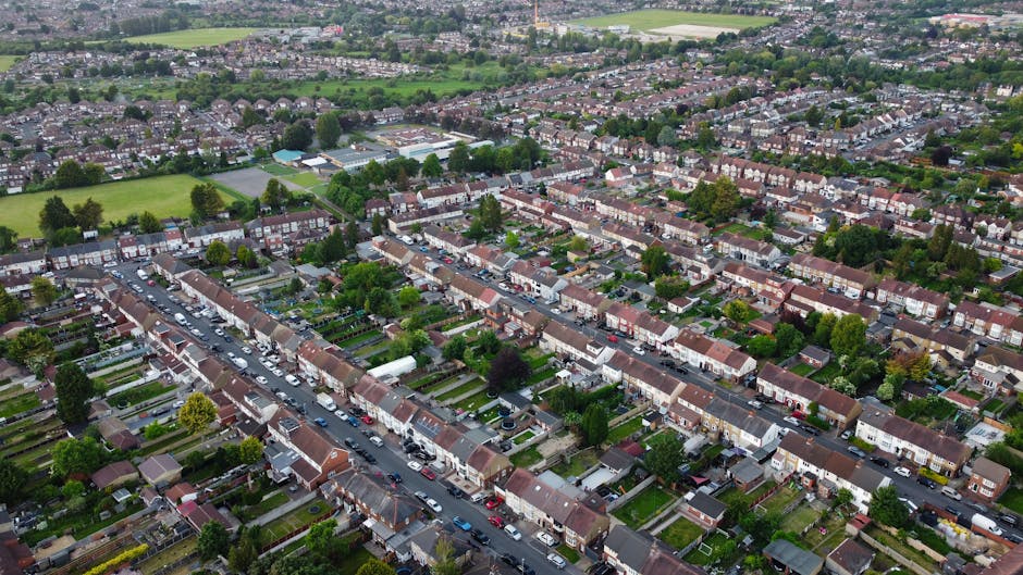 An aerial view of a residential neighbourhood showing multiple rows of terraced houses with brick facades and small front gardens, situated along narrow streets filled with parked cars and driveways. The image captures the rooftops, greenery, and trees interspersed within the area, with some open green spaces and sports fields visible in the background. The scene depicts a typical urban area suitable for house removals and home relocation services. The natural daylight illuminates the scene clearly, highlighting the uniformity and orderly layout of the housing. This type of setting often requires professional moving companies like Man with Van Canonbury to assist with furniture transport, packing, and loading process during urgent eviction moves or planned relocations.