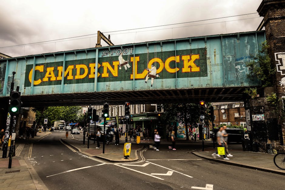 A large elevated railway bridge in an urban setting featuring a weathered turquoise-green metal structure with the words 'Camden Lock' painted in bold yellow letters, accompanied by painted figures of children hanging from the bridge. The area beneath the bridge shows a street crossing with traffic lights, and pedestrians walking along the pavement. There are vehicles and delivery trucks on the road, with some people blurred due to motion. The surroundings include buildings with brick facades and graffiti, and some greenery is visible nearby. This scene captures a lively environment typical for a busy London neighbourhood, with potential relevance to house removals or moving logistics in the Camden area, as managed by companies like Man with Van Canonbury.